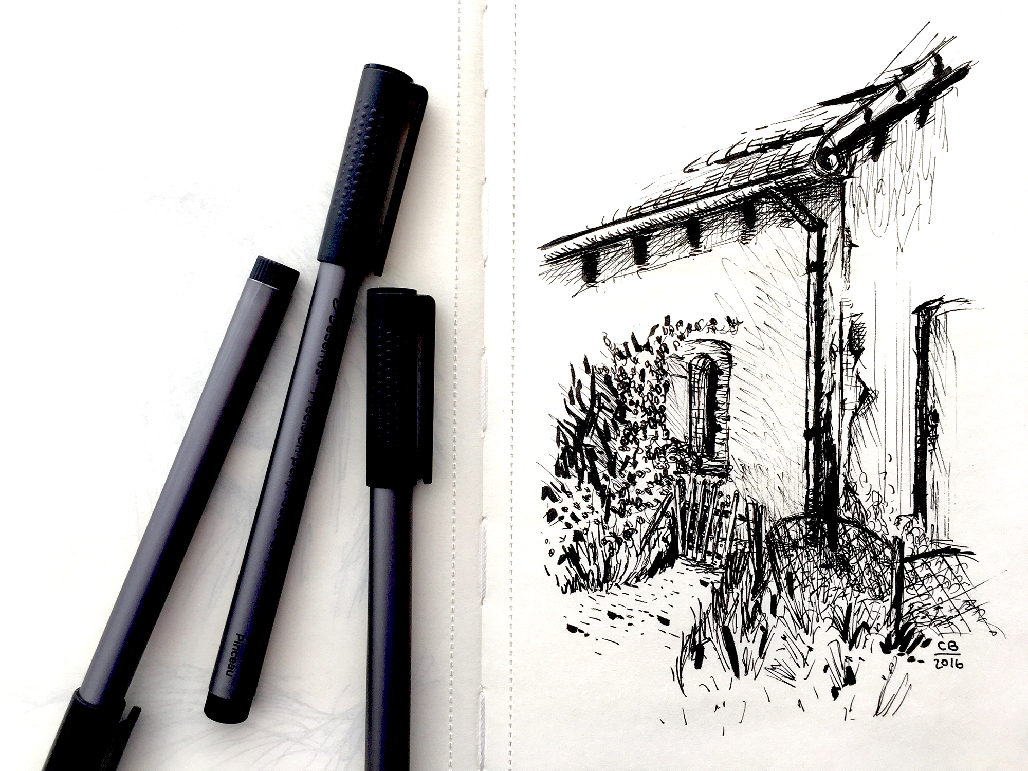 A sketchbook open to a pen sketch of the corner of a house surrounded by garden features, three pens resting on the left page of the book.