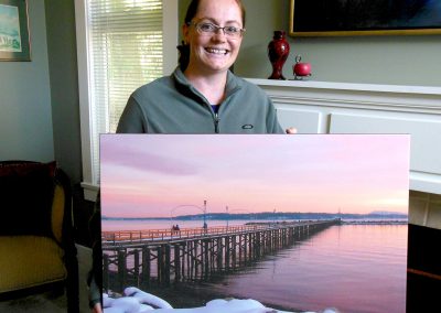 Cheryl poses with a canvas-printed photograph of a pier in a pink winter sunset